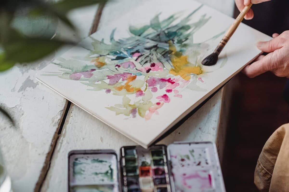 There is a watercolour painting of flowers in bright colours with a paintbrush being held by a person. In the foreground is half a watercolour palette.