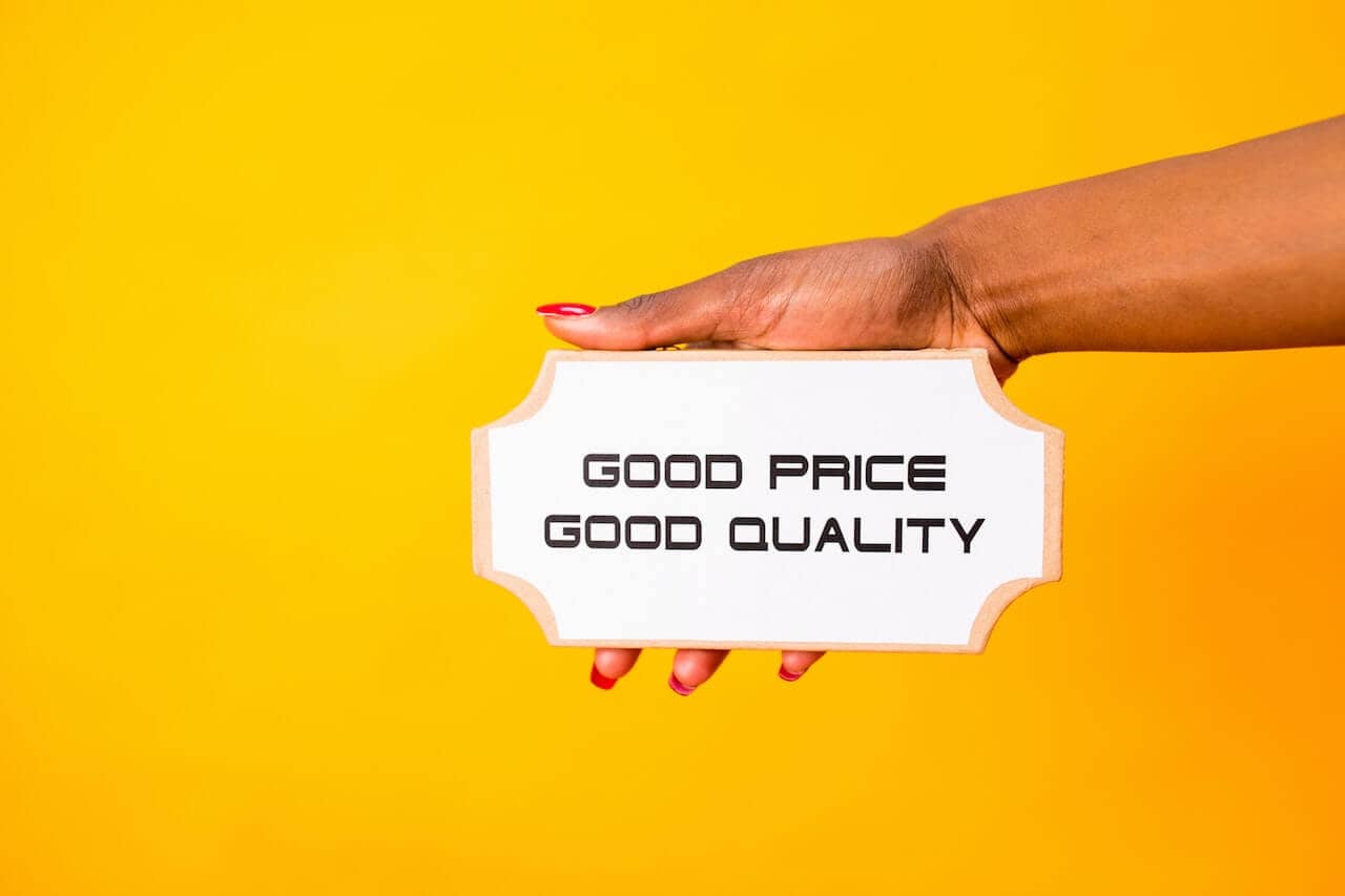 Yellow background with a person of colours hand and arm reaching in from the right. Holding a sign stating 'Good price, good quality' on a white background with black writing.