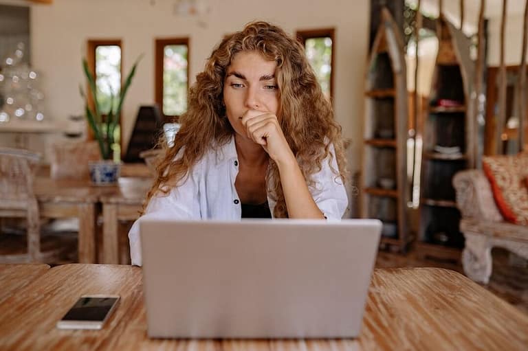 Caucasian woman with long, brown, wavy hair is sitting at a timber desk thinking while looking at a laptop.